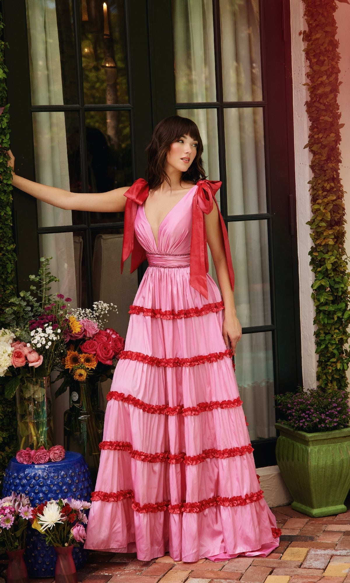 A woman wears the Ava Presley 40091 Shoulder Bows A-Line Formal Dress in pink and red, standing before glass doors with potted plants and floral arrangements around her.